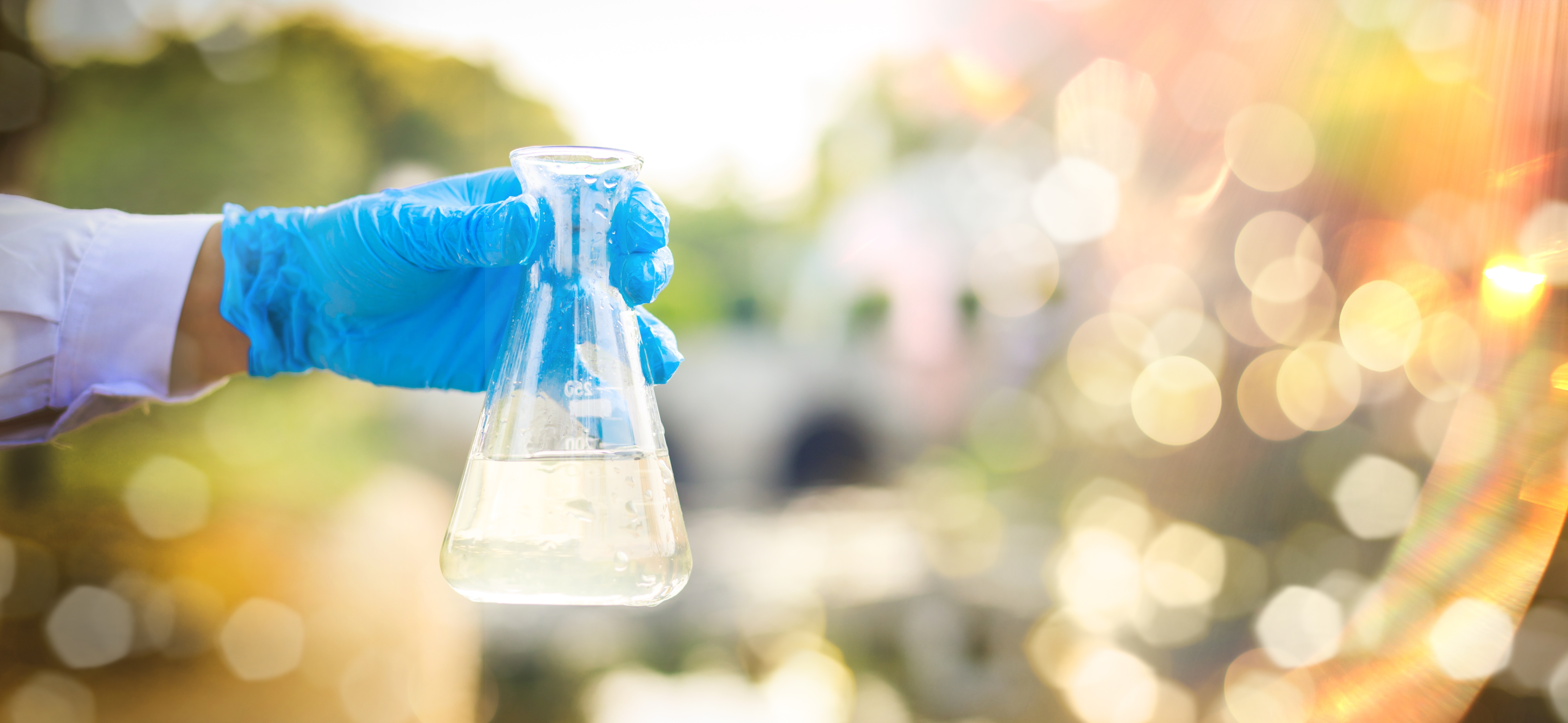 An image od a scientist holding a lab beaker with blue gloves on; the scientist is standing outside with natural light.